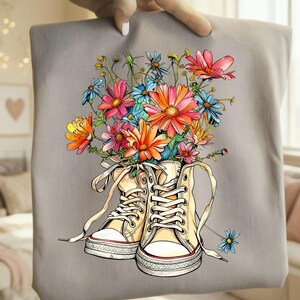 May include: A gray tote bag featuring a colorful floral design. The artwork depicts a pair of cream-colored high-top sneakers filled with a vibrant bouquet of pink, orange, and blue flowers. The bag is held by a hand.
