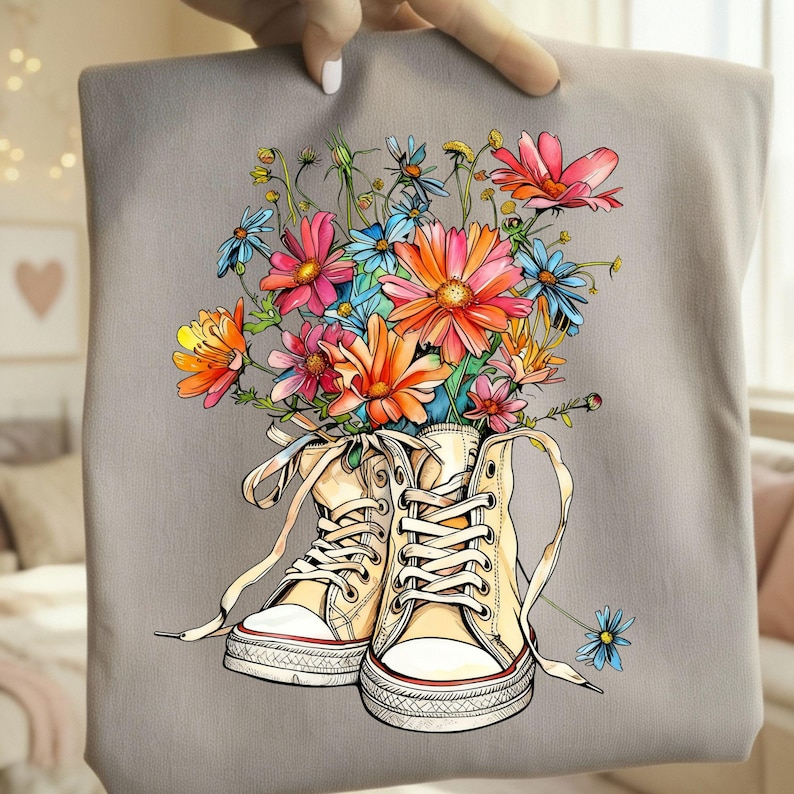 May include: A gray tote bag featuring a colorful floral design. The artwork depicts a pair of cream-colored high-top sneakers filled with a vibrant bouquet of pink, orange, and blue flowers. The bag is held by a hand.