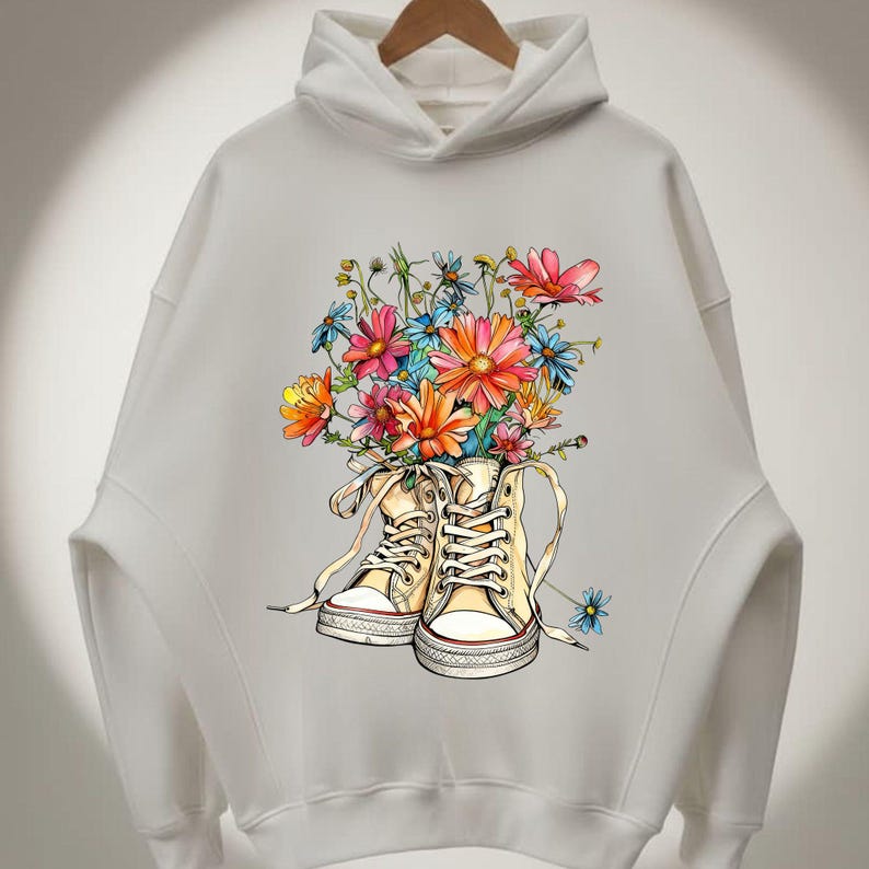 May include: White hoodie with a colorful floral design. The graphic features a pair of cream-colored high-top sneakers filled with a bouquet of pink, orange, and blue flowers. The hoodie has a drawstring hood and long sleeves.
