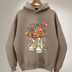 May include: A tan hoodie featuring a graphic of high-top sneakers filled with colorful flowers. The sneakers are cream-colored with white laces and a red stripe on the sole. The flowers are a mix of pink, orange, blue, and yellow.