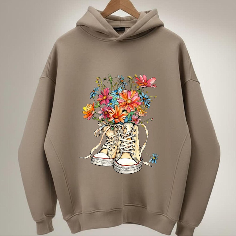 May include: A tan hoodie featuring a graphic of high-top sneakers filled with colorful flowers. The sneakers are cream-colored with white laces and a red stripe on the sole. The flowers are a mix of pink, orange, blue, and yellow.