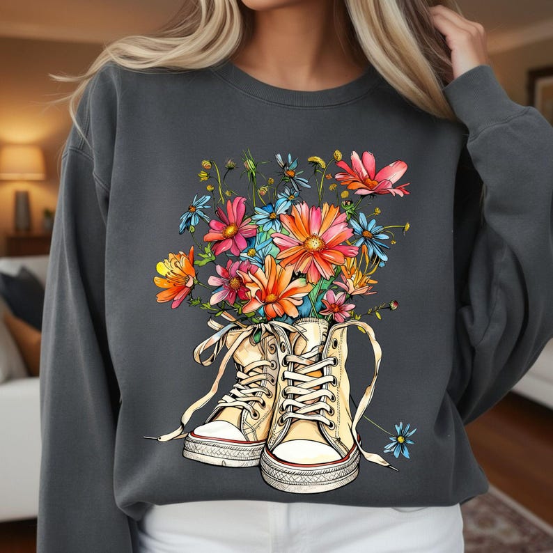 May include: Dark gray sweatshirt featuring a colorful floral design. The graphic shows a pair of cream-colored high-top sneakers filled with vibrant flowers in shades of pink, orange, and blue. The sweatshirt has a crew neck and long sleeves.