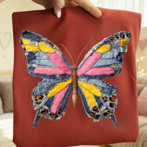Puede incluir: Un cojín cuadrado de color óxido con un gran diseño de mariposa. La mariposa está hecha de mezclilla, tela amarilla, rosa y con estampado de leopardo, con una apariencia texturizada y cosida. El cuerpo de la mariposa es de material marrón tejido.
