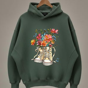 May include: Dark green hoodie featuring a colorful floral design. The graphic depicts a pair of cream-colored high-top sneakers overflowing with vibrant flowers in shades of pink, orange, blue, and yellow. The hoodie has a hood and long sleeves.