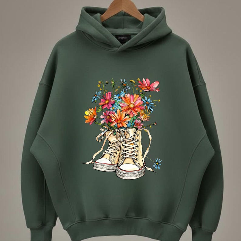 May include: Dark green hoodie featuring a colorful floral design. The graphic depicts a pair of cream-colored high-top sneakers overflowing with vibrant flowers in shades of pink, orange, blue, and yellow. The hoodie has a hood and long sleeves.