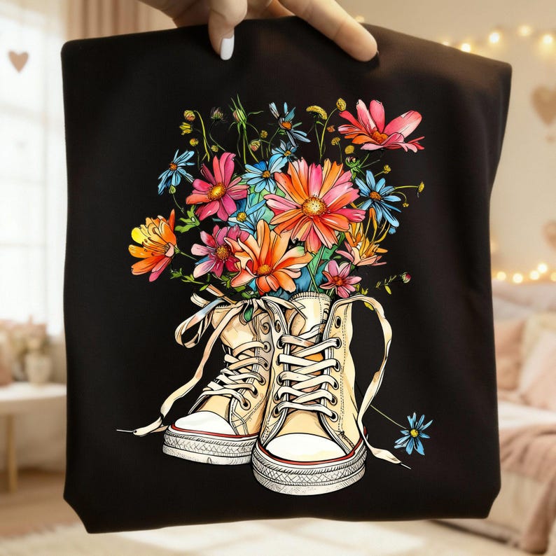 May include: Black tote bag featuring a colorful floral design. The artwork depicts a pair of cream-colored high-top sneakers overflowing with vibrant flowers in shades of pink, orange, and blue. The bag is held by a hand.