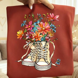 May include: A maroon tote bag featuring a vibrant illustration of cream-colored sneakers overflowing with colorful flowers. The flowers include pink, orange, blue, and yellow blooms. The sneakers have white laces and a red stripe on the sole.