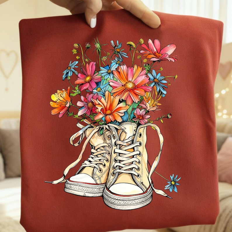 May include: A maroon tote bag featuring a vibrant illustration of cream-colored sneakers overflowing with colorful flowers. The flowers include pink, orange, blue, and yellow blooms. The sneakers have white laces and a red stripe on the sole.