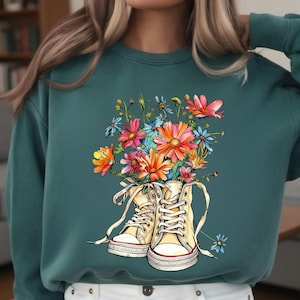 May include: Teal sweatshirt featuring a graphic of high-top sneakers filled with colorful flowers. The sneakers are cream-colored with white laces and red accents. The flowers include pink, orange, blue, and yellow blooms.