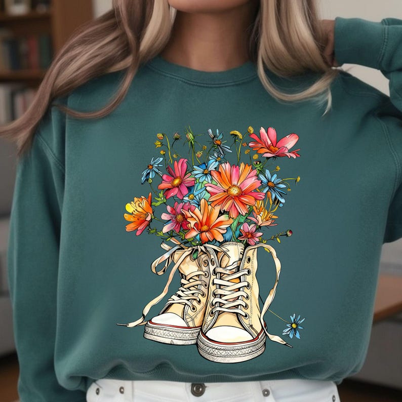 May include: Teal sweatshirt featuring a graphic of high-top sneakers filled with colorful flowers. The sneakers are cream-colored with white laces and red accents. The flowers include pink, orange, blue, and yellow blooms.