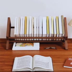 Op de afbeelding: Een houten boekenplank toont een verzameling boeken met witte en gele ruggen. Een open boek, een bril en een klein notitieboekje liggen op het houten oppervlak. De boekenplank heeft een warme bruine kleur.