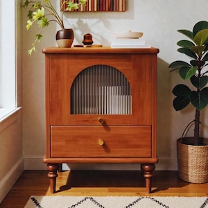 May include: A wooden cabinet in a warm, reddish-brown hue. It has a curved glass door with vertical lines and a drawer with a brass knob. The cabinet stands on short, turned legs. A plant and decorative items are on top.