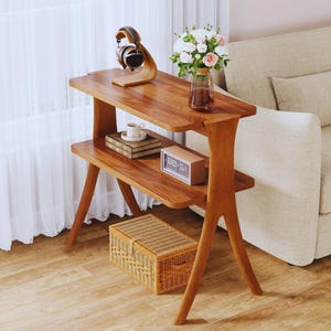 May include: A wooden side table with two shelves, holding a pair of headphones, a vase of flowers, a small clock, and a decorative box. The table has a natural wood finish and a unique leg design. A woven basket sits on the floor below.