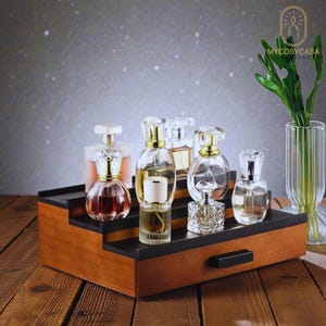 May include: A wooden display stand with steps, holding six perfume bottles. The bottles are clear glass with gold or silver lids. The stand has a black top and a small drawer. A vase with green leaves is on the right.