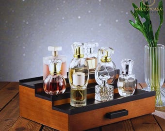 May include: A tiered wooden display stand with six perfume bottles of various shapes and sizes. The bottles are clear glass with gold or silver accents. The stand has a black top and a small drawer. A vase with green leaves is on the right.