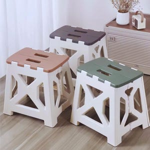 May include: Three folding step stools in the image. Each stool has a white frame and a colored top. The tops are brown, dark gray, and green. The stools are made of plastic and have a geometric design.