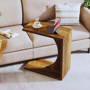 May include: A wooden C-shaped side table with a warm brown finish. The table has a curved design on one side and a flat top. A laptop, coffee cup, and magazine rest on the table. The table is next to a beige sofa.