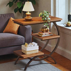 Può includere: Tavolino a tre livelli con struttura in metallo marrone scuro e ripiani in legno. Il ripiano superiore contiene una lampada con paralume beige. Il ripiano centrale ha libri e un elefante decorativo. Il ripiano inferiore ha un cestino.