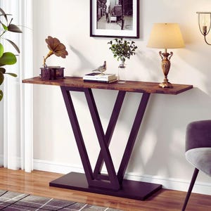 Può includere: Una consolle marrone con una base a forma di V in metallo scuro. Il piano del tavolo è in legno e ha un aspetto vintage. Un grammofono, una lampada e altri oggetti decorativi sono sul tavolo. Il tavolo è appoggiato a un muro bianco.