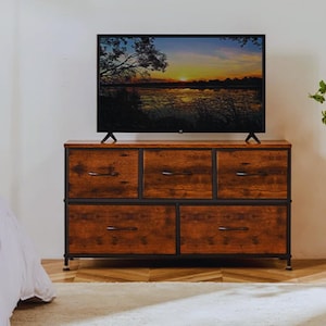 May include: A wooden TV stand with five drawers and a black metal frame. The drawers have a rustic, distressed wood finish. A flat-screen TV displays a sunset scene. The stand is on a light-colored rug.
