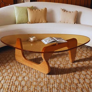 May include: A mid-century modern coffee table with a clear, oval-shaped glass top and a wooden base. The base is made of two curved wooden supports. A book and a teacup sit on the table. The table is on a woven rug.