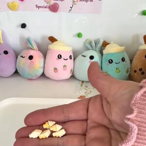 Può includere: Giocattoli di peluche a forma di bubble tea e coniglietti in tonalità pastello. Una mano tiene in mano diversi piccoli ciondoli a forma di croissant color crema. È visibile un elenco di accessori.