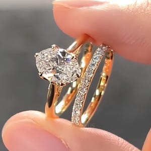 Op de afbeelding: Een verlovingsringset van gepolijst geelgoud, bestaande uit een ring met een grote ovaal geslepen middensteen in een klauwzetting en een bijpassende band bezet met kleine ronde diamanten, wat zorgt voor een schitterende en elegante uitstraling.
