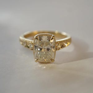 May include: A gold engagement ring featuring a large, rectangular-cushion cut diamond. The band is gold and adorned with smaller diamonds. The ring is set against a soft, neutral background. The text "BEZELHAUS" is visible on the ring.