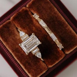 May include: Two gold rings with clear gemstones displayed in a burgundy velvet ring box. The engagement ring features an emerald-cut center stone flanked by pear-shaped stones. The wedding band has rectangular stones.