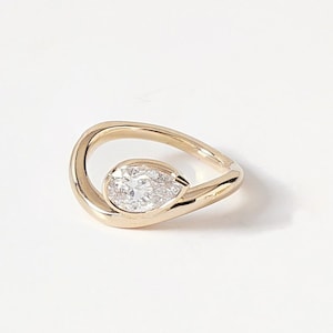 Puede incluir: Un anillo de oro con una gema clara en forma de pera, engastada en un dise&ntilde;o abierto y fluido. La banda del anillo se curva alrededor de la gema, creando un aspecto moderno. El anillo est&aacute; sobre un fondo blanco.