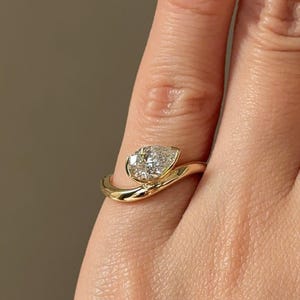 Puede incluir: Un anillo de oro con un diamante en forma de pera. El diamante est&aacute; engastado en un bisel, y la banda se curva alrededor del dedo. Una joya delicada.
