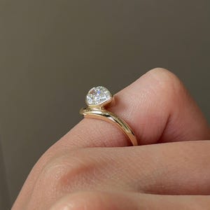 Puede incluir: Un anillo de oro con una gema grande y transparente en forma de coraz&oacute;n. La banda del anillo se curva alrededor del dedo, creando un dise&ntilde;o &uacute;nico y elegante. La gema est&aacute; engastada en un bisel. El anillo se muestra en un dedo.