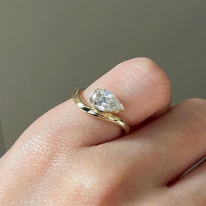 Puede incluir: Un anillo de oro con un diamante en forma de pera. El diamante est&aacute; engastado en un bisel y la banda se curva alrededor del dedo. El anillo se muestra en el dedo de una persona.