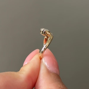 Puede incluir: Un anillo de oro con una piedra central de diamante y un dise&ntilde;o curvo &uacute;nico. El anillo se sostiene, mostrando sus delicados detalles y el brillo del diamante. La banda del anillo tiene una apariencia retorcida.