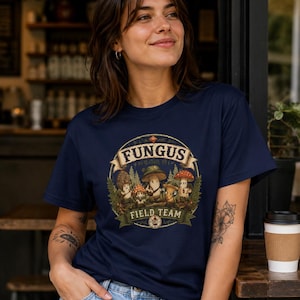 May include: Navy blue t-shirt featuring a circular graphic with the text "FUNGUS FIELD TEAM." The graphic depicts various cartoon mushrooms in a forest setting. The shirt is made of a soft material.