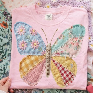 Puede incluir: Camiseta rosa claro con un gran diseño de mariposa patchwork. Las alas de la mariposa están compuestas por varias telas estampadas en tonos azules, amarillos y rosas, con detalles florales bordados. La camiseta tiene cuello redondo y mangas cortas.