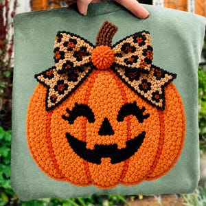 Puede incluir: Una sudadera verde salvia con una gran aplicación de calabaza naranja texturizada y una cara de jack-o'-lantern cosida en negro. La calabaza tiene un tallo marrón y un gran lazo decorativo con un patrón de estampado de leopardo beige y negro.