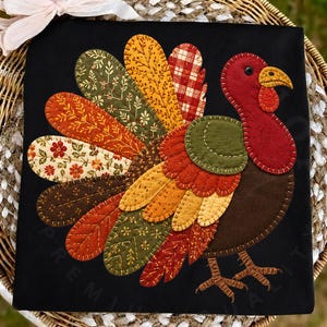 May include: A black t-shirt featuring a large, colourful turkey appliqué made from various fabric patches. The turkey has a red head, a brown body, and a fan-shaped tail composed of patterned leaves and feathers in shades of orange, yellow, green, and red.