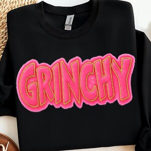 May include: Black sweatshirt with the word "GRINCHY" in large, pink, fuzzy letters outlined in gold. The sweatshirt has a crew neck and long sleeves. The word "GRINCHY" is the focal point of the design.