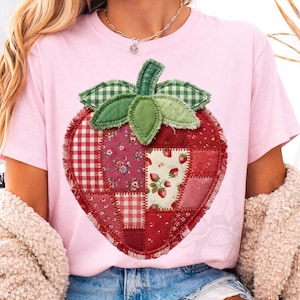 Puede incluir: Camiseta rosa claro con un gran diseño de fresa en patchwork. La fresa está compuesta por varias telas rojas y rosas, incluyendo cuadros y estampados florales. Hojas verdes y a cuadros coronan la fresa.