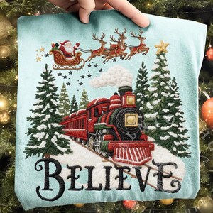 Puede incluir: Un cojín azul claro con un diseño bordado navideño. El diseño presenta a Papá Noel en un trineo tirado por renos, un tren rojo, árboles de Navidad y la palabra "BELIEVE" en escritura negra.
