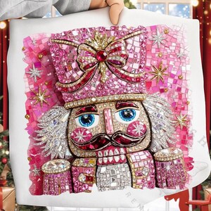 May include: A decorative pillow featuring a pink and silver mosaic design of a nutcracker soldier. The nutcracker has blue eyes, a red mustache, and is adorned with a crown and bow. The pillow is held by a person.