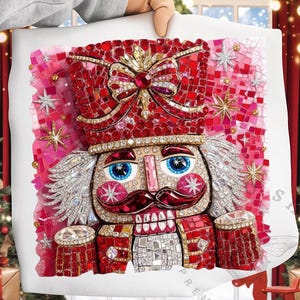 Könnte beinhalten: Ein dekoratives Kissen mit einem Mosaikbild eines Nussknacker-Soldaten. Der Nussknacker hat einen roten Hut mit einer Schleife, einen Schnurrbart und blaue Augen. Das Kissen hat einen rosa und roten Hintergrund mit Sternakzenten. Das Kissen wird von einer Person gehalten.