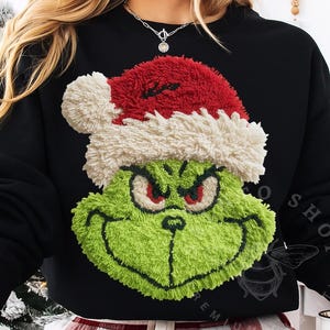 May include: Black sweatshirt with a green Grinch face wearing a red and white Santa hat. The Grinch's face has a textured appearance with red eyes and a grin. A festive holiday apparel item.