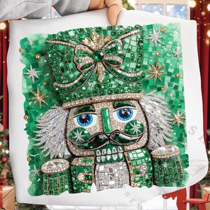 May include: A cushion with a mosaic-style nutcracker design. The nutcracker is decorated with green, silver, and gold embellishments, including a hat, bow, and star accents. The eyes are blue, and the overall theme is festive.