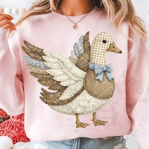 May include: Pale pink sweatshirt with a patchwork duck illustration. The duck has a gingham head, a blue bow, and wings made of patterned fabrics in brown, blue, and cream. The sweatshirt is a soft pink colour.