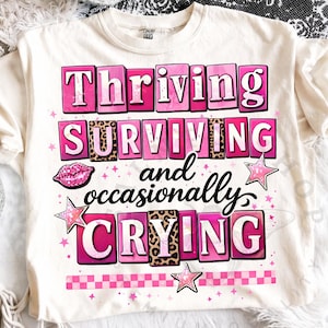 May include: Cream-colored t-shirt with the text "Thriving, Surviving, and Occasionally Crying" in pink and leopard print lettering. The design includes a pink lip graphic and star accents.