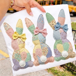 May include: White fabric featuring three patchwork bunny rabbits with colorful floral and patterned designs. Each bunny has a bow and a fluffy white tail. The bunnies are in pastel colors, perfect for Easter.