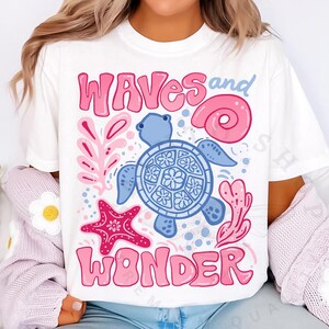 Pu&ograve; includere: T-shirt bianca con un design grafico colorato raffigurante una tartaruga blu, una stella marina rosa e le parole "Waves and Wonder" in rosa. Il design include elementi a tema marino come una conchiglia e coralli.