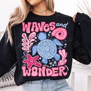 Pu&ograve; includere: Felpa nera con un design grafico colorato. Il design presenta una tartaruga blu, una stella marina rosa e le parole "WAVES and WONDER" in lettere rosa. La felpa &egrave; a girocollo.
